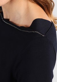 Marinblå off-shoulder topp i ett slätt tyg med metallisk kant runt halsringningen. Enkel design med en figurnära silhuett.