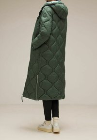 Abrigo acolchado verde con un patrón de rombos, que incluye capucha, bolsillos laterales y lados con cremallera. Llevado sobre pantalones negros y zapatillas.