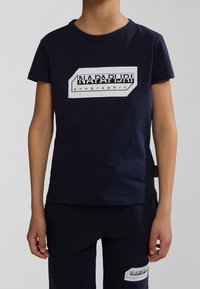 T-paita tummansinisestä puuvillasta, jossa on valkoinen graafinen logo, jossa on tekstit "NAPAPIJRI" ja "geographic" edessä; lyhyet hihat ja pyöreä kaula-aukko.