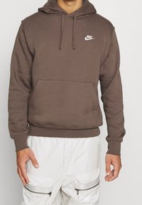 Brun huvtröja i mjukt material, med en framficka, ribbade muddarna och en vit Nike-logga på bröstet.