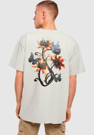 Lys beige oversized T-skjorte med livlige blomster- og sommerfugltrykk i oransje, svart og grå, med tekst på baksiden.