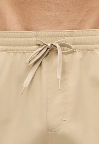Close-up af beige elastiske shorts med bundet snøre i taljen på en persons talje med synlig hud over.