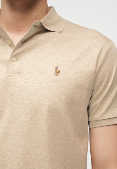 Μπεζ πόλο πουκάμισο με γιακά και δύο κουμπιά, με μικρό κεντημένο λογότυπο παίκτη πόλο πάνω σε άλογο στο αριστερό μέρος του στήθους.