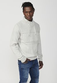Grau strukturierter Pullover mit hohem Kragen und Kordelzug. Mit einem auffälligen, geprägten Logo in der Mitte. Kombiniert mit blauen, ausgewaschenen Jeans.