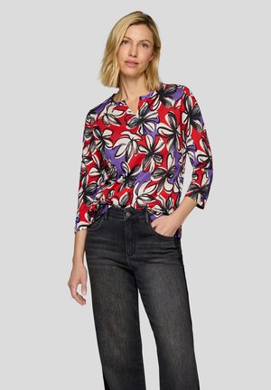 Bunt gemusterte Bluse mit einem Design in Rot, Lila und Schwarz, V-Ausschnitt und drei Viertelärmel, kombiniert mit dunkelblauen Jeans.