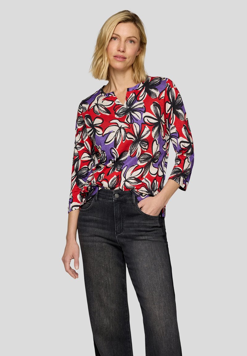 Bunt gemusterte Bluse mit einem Design in Rot, Lila und Schwarz, V-Ausschnitt und drei Viertelärmel, kombiniert mit dunkelblauen Jeans.