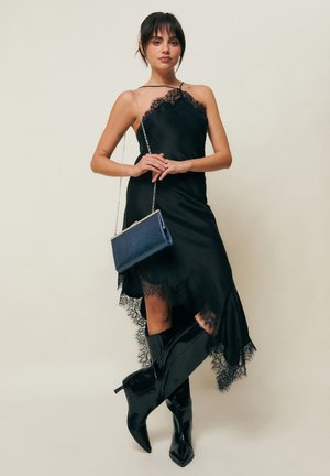 Naine kannab asümmeetrilist musta pitsiga kaunistatud kleiti, musti lakknahast põlveni saapaid ning hoiab kandikul ainet sinist kandekotti kettkinnitusega.