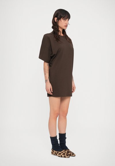 Γυναίκα με μακριά σκούρα μαλλιά, φοράει χαλαρό καφέ φόρεμα με κοντά μανίκια, σκούρες μπλε κάλτσες και φλατ παπούτσια με λεοπάρ σχέδιο, στέκεται πλαγίως.