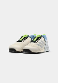 Des baskets avec une tige en mesh beige clair, une semelle en caoutchouc noire, des accents verts et bleus, et trois bandes diagonales sur le côté.