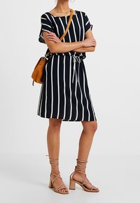 Mekko, jossa on sinivalkoiset raidat, lyhyet hihat, kiristettävä vyötärö ja polven pituinen helma. Yhdistetty beigenväristen paksupohjaisten sandaalien ja ruskean laukun kanssa.