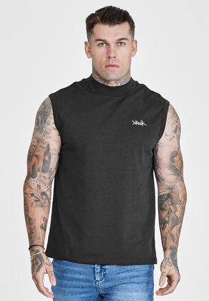 Mann med tatoveringer har på seg en svart ermeløs topp med liten hvit logo på brystet og blå jeans, vendt mot kamera mot en ensfarget bakgrunn.