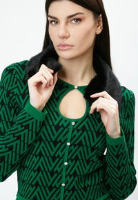 Grüner und schwarzer gemusterter Cardigan mit einem Schlüsselloch-Ausschnitt, Kragen aus Kunstfell und dekorativen Knöpfen entlang der Vorderseite. Weiche Textur.