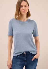 Junge Frau mit schulterlangem blonden Haar, die ein hellblaues Kurzarm-Strickoberteil und dunkelblaue Jeans trägt, mit einer Hand in der Tasche.