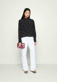 Blusa preta com bolinhas coloridas, calças largas brancas e uma carteira metálica rosa. Modelo em pé com sandálias pretas sobre um fundo neutro.