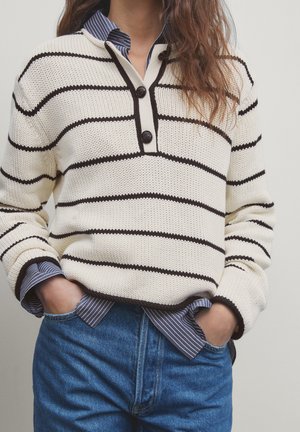 Persoon in een roomkleurige en zwarte gestreepte gebreide trui over een blauw-witte gestreepte shirt, gestoken in hoge taille blauwe jeans met de handen in de zakken.