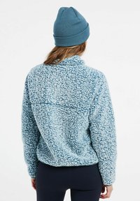 Pullover in pile azzurro chiaro con colletto alto, superficie testurizzata e vestibilità rilassata. Un berretto blu è indossato sulla testa.