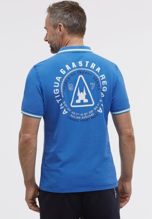 Mann mit kurzen Haaren und Bart, trägt ein blaues Poloshirt mit weißem und hellblauem Besatz, das ein nautisch inspiriertes Logo und Text auf der Rückseite zeigt.