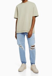 Man som bär en överdimensionerad beige t-shirt, ljusblåa slitna jeans med elastiska manschetter och beige sneakers, står mot en vit bakgrund.