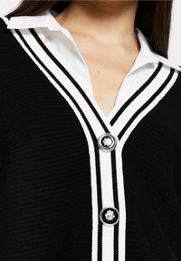Zwarte gebreide cardigan met een V-hals, witte gestreepte accenten en twee ronde knopen met een contrasterend ontwerp bij de sluiting.