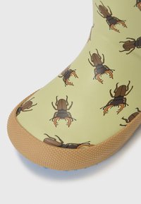 Botte en caoutchouc avec une surface vert clair, ornée de motifs répétés de coccinelles marron foncé. Accentuée d'une semelle en tan texturée et d'un intérieur bleu.