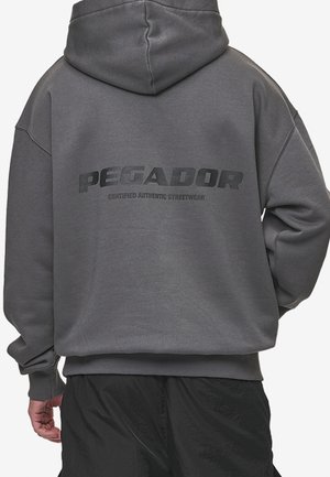 Mann trägt dunkelgrauen Hoodie mit dem Schriftzug „PEGADOR Certified Authentic Streetwear“ auf dem Rücken, kombiniert mit schwarzen Hosen.