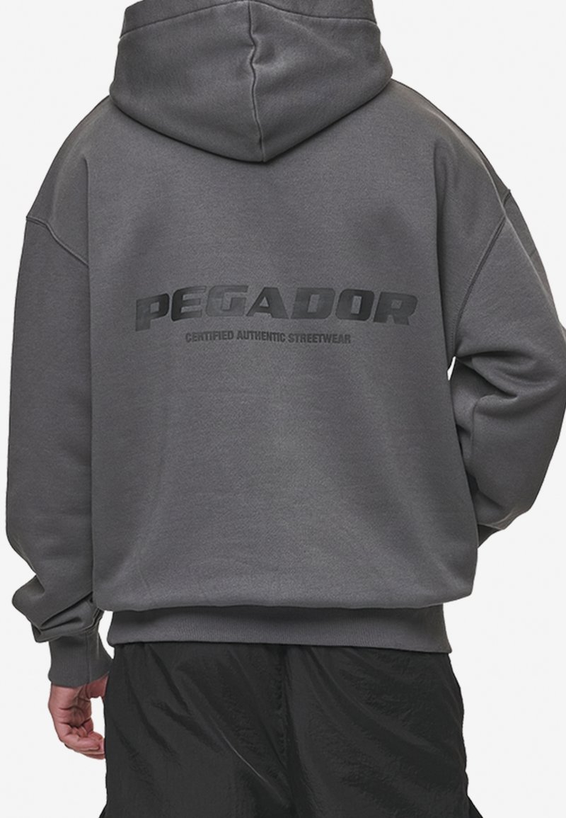 Homme portant un sweat à capuche gris foncé avec le texte « PEGADOR Certified Authentic Streetwear » dans le dos, assorti à un pantalon noir.
