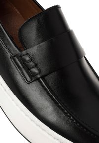 Nahaufnahme eines schwarzen Lederloafers mit breitem Riemen und weißer Gummisohle, die detaillierte Nähte und ein braunes Innenfutter zeigt.