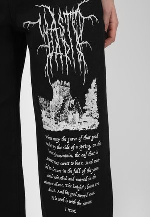 Pantalon noir avec texte blanc de style gothique, un graphique de château médiéval en pierre et un vers poétique imprimé sur la cuisse.