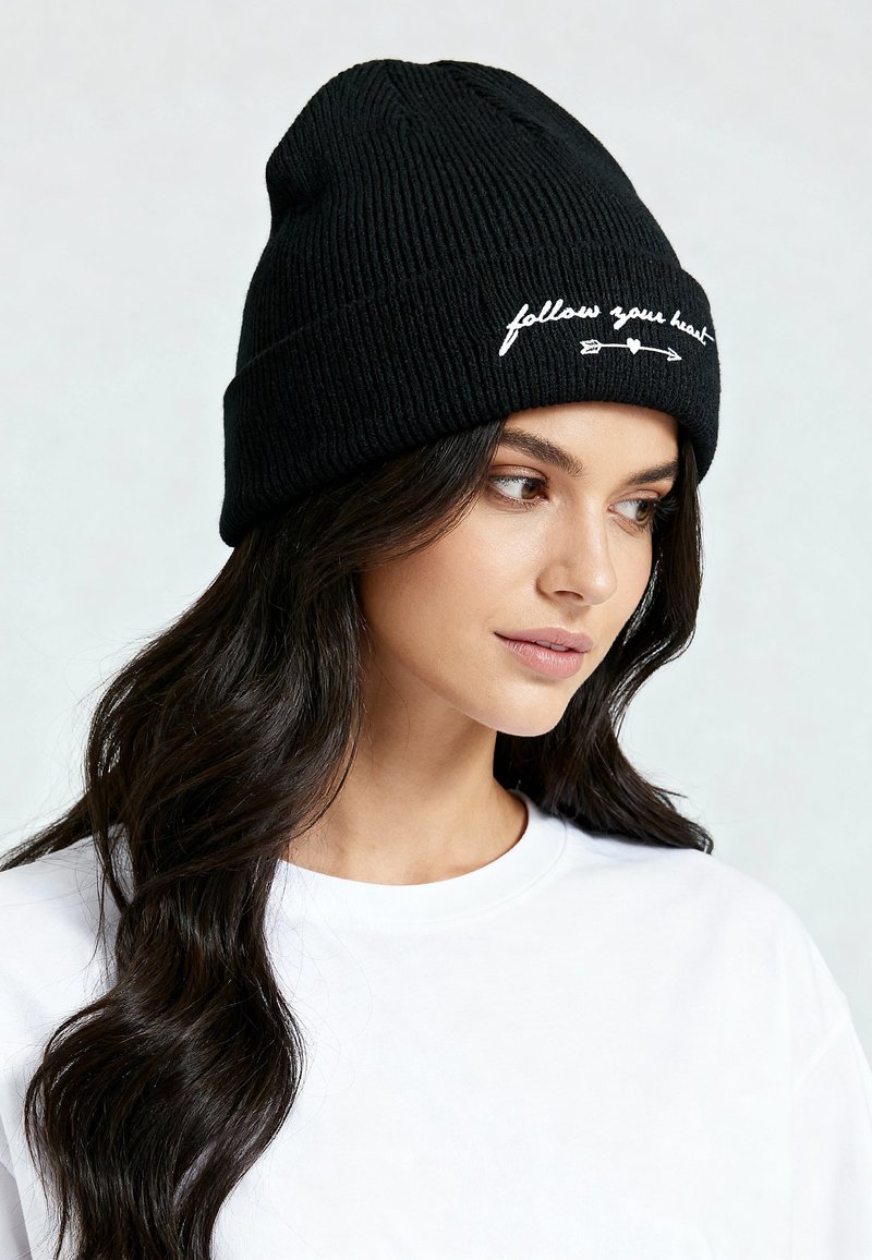 Bonnet en tricot côtelé noir avec texte brodé blanc « follow your heart », au design décontracté. Le texte est centré sur le devant.