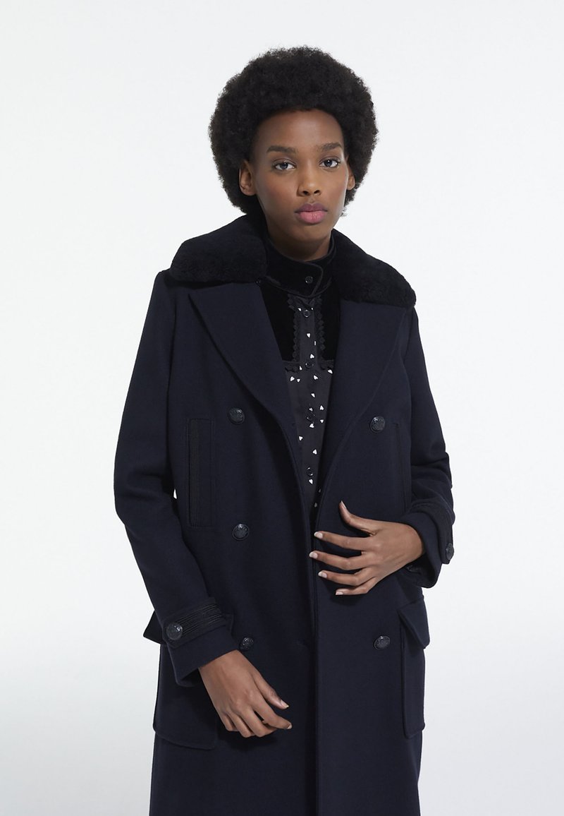 Manteau en laine bleu marine avec un design croisé, grand col noir et poches latérales. Caractérisé par un tissu texturé et des boutons proéminents.