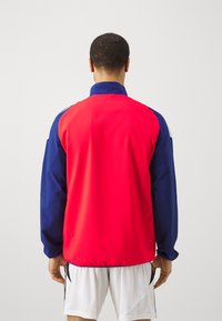 Chaqueta cortaviento roja y marina con un alto cuello, puños elásticos y una textura suave, que presenta un diseño a bloques de color.