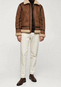 Man in een bruin schapenvachten jack met zwarte accenten, crèmekleurige broek, donkerbruin coltrui en donkerbruine leren laarzen.