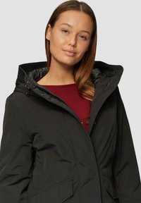 Schwarze kapuzenparka mit Reißverschluss und Druckknopfverschluss, ausgestattet mit einem gesteppten Innenfutter und Fronttaschen, getragen über einem burgunderfarbenen Oberteil.