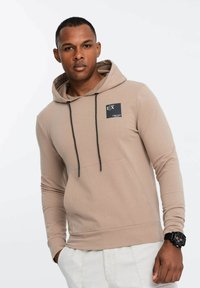 Felpa beige con cappuccio, cordini neri, tasca a marsupio e una piccola etichetta con logo. Realizzata in tessuto morbido per una vestibilità comoda.