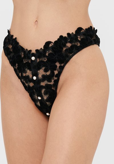 String tanga en dentelle florale noire avec tissu transparent, présentant des fleurs texturées, une taille haute et des boutons décoratifs en perles sur le devant.
