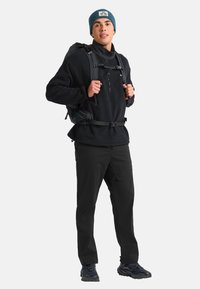 Zwarte fleece pullover met halve rits, zwarte broek, blauwe gebreide beanie en een zwarte rugzak. Stevige schoenen met textuurzolen.