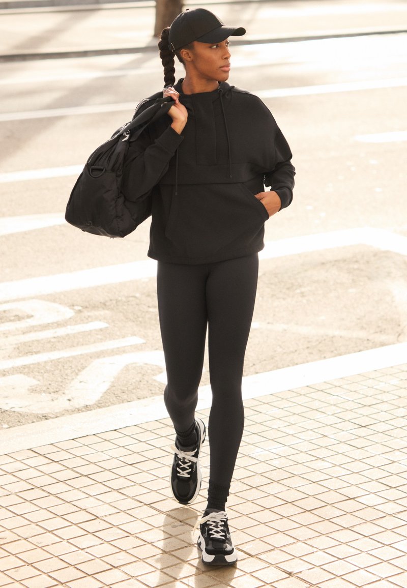 Svart idrottshoodie, åtsittande svarta leggings och svarta sneakers med vita accenter. Bär en svart basebollkeps och har en svart ryggsäck.