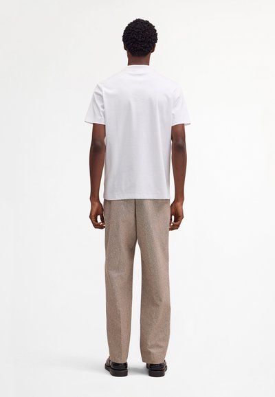 Man with short curly hair stands facing away, wearing a plain white t-shirt, beige textured pants, and black shoes on a white background.