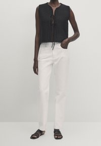 Gilet nero corto in maglia con chiusura a fiocco, abbinato a jeans bianchi dritti. Il modello indossa sandali neri intrecciati. Design e texture minimalisti.