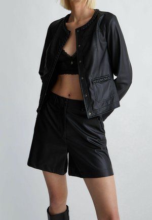 Femme portant une veste en cuir noire avec bordure tressée, un short en cuir noir, un bralette en dentelle noire et des bottes noires sur fond uni.