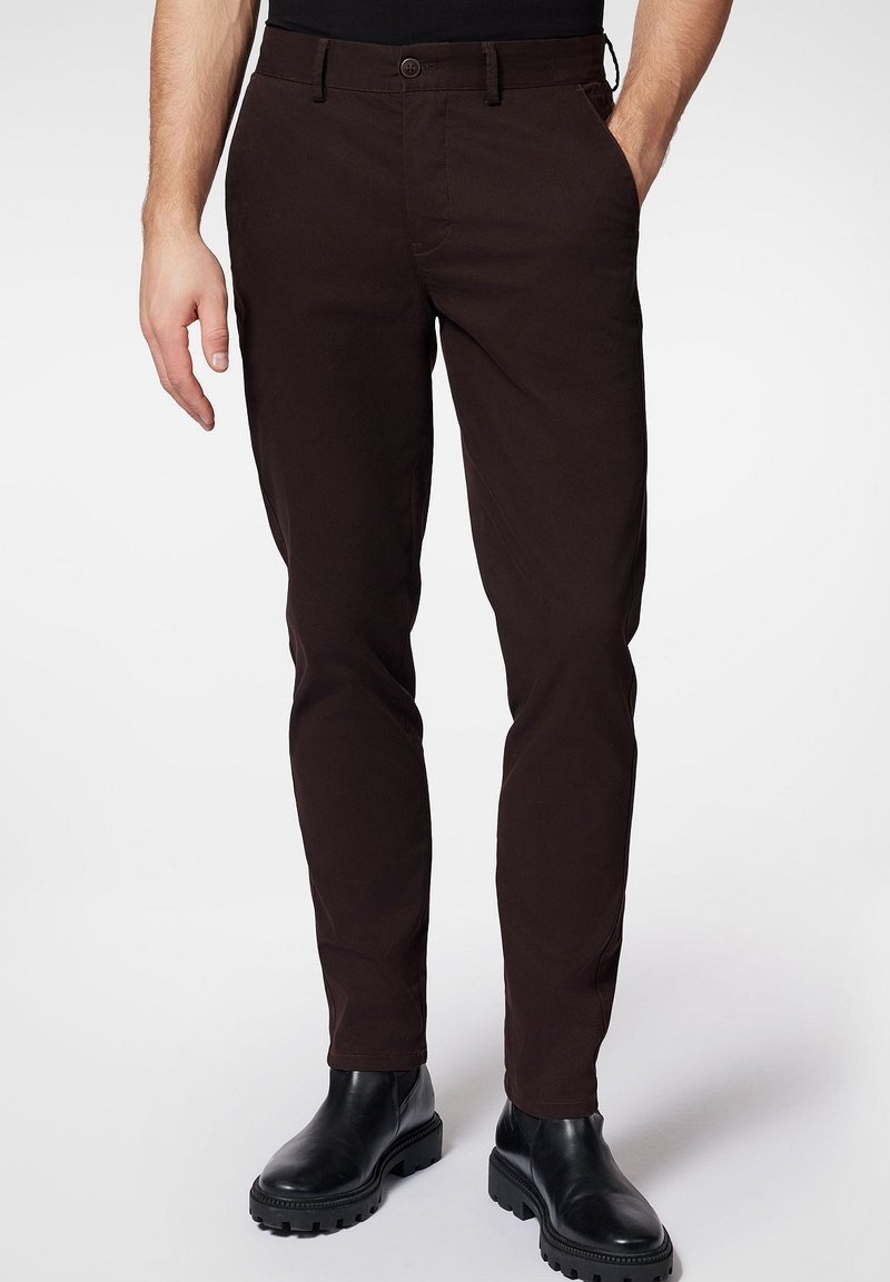 Homme portant un pantalon slim marron foncé avec des poches latérales et des bottes en cuir noir, debout avec une main dans la poche.