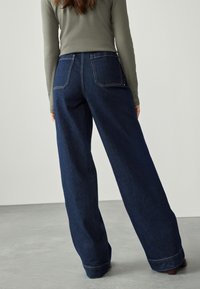 Jeans a gamba larga in denim blu scuro con vita alta, tasche posteriori visibili e cuciture a contrasto. Texture liscia con una leggera lucentezza.