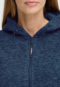 Jūras zila kapuce ar rāvējslēdzēju, izgatavota no teksturēta auduma. Tam ir kapuce ar regulējamiem sašūtiem auklām un priekšējais rāvējslēdzējs ar zīmola nosaukumu.