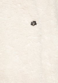 Hvid kunstig pels med en blød, fluffet tekstur og en lille sort metalknapdetalje. Overfladen viser små variationer i fiberen.