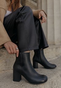 Botine negre din piele cu vârf rotund și toc pătrat, având o textură netedă și cusături subtile, asortate cu pantaloni din piele sintetică negri.