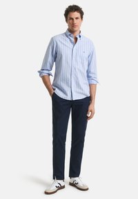 Chemise boutonnée à rayures bleu clair avec des manches longues, associée à un pantalon bleu marine et des baskets blanches avec des accents marron. Design simple, coupe décontractée.