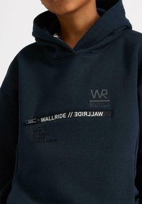 Granatowa bluza z kapturem z logo i graficzną taśmą z napisem "WALLRIDE". Miękka tkanina o luźnym kroju. Zawiera nazwy miast w kolorze czarnym.