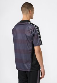 Hombre vistiendo una camiseta deportiva de rayas oscuras con cinta de logo en las mangas, visto desde atrás contra un fondo blanco liso.