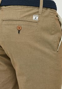 Tan textured pants feature a back pocket with a brown button closure and a white label with "INDICODE PREMIUM GOODS." Woven navy belt visible.
