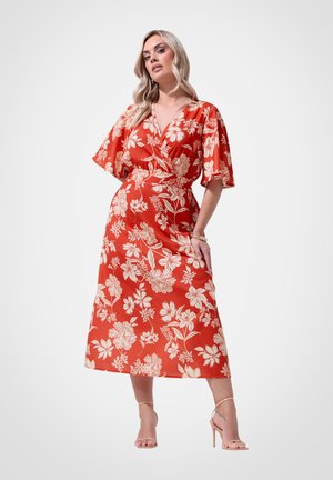 Kobieta w czerwonej sukience midi z białym kwiatowym wzorem, z falbanianymi rękawami, dekoltem w serek oraz beżowymi sandałami na wysokim obcasie, pozująca z jedną ręką na biodrze.
